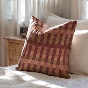 Pu&ograve; includere: Un cuscino quadrato con un motivo in tessuto intrecciato a strisce verticali alternate nei toni del bordeaux intenso, verde oliva tenue e rosa polvere delicato. Il tessuto presenta una trama visibile e un design geometrico a blocchi costante.