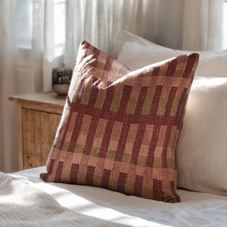 Pu&ograve; includere: Un cuscino quadrato con un motivo in tessuto intrecciato a strisce verticali alternate nei toni del bordeaux intenso, verde oliva tenue e rosa polvere delicato. Il tessuto presenta una trama visibile e un design geometrico a blocchi costante.