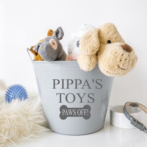 Könnte beinhalten: Ein hellgrauer Metalleimer mit der Aufschrift PIPPA'S TOYS und PAWS OFF! in einer knochenförmigen Grafik. Der Eimer ist mit verschiedenen Plüschtieren gefüllt, darunter ein beiger Hund, ein grauer Esel und ein weißes Spielzeug, mit einem blauen Ball.