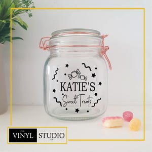 Puede incluir: Un tarro de cristal transparente con tapa abisagrada y cierre color cobre. El tarro está decorado con texto y gráficos negros que dicen "KATIE'S Sweet Treats". Varios caramelos son visibles frente al tarro. El logotipo de Vinyl Studio GB está en la esquina inferior izquierda.