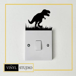 May include: A black Tyrannosaurus Rex dinosaur silhouette decal, standing on grass, is affixed above a white light switch. The decal is a decorative element for a wall. The Vinyl Studio GB logo is in the lower left corner.