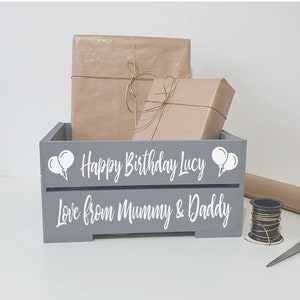 May include: A gray wooden crate with the text "Happy Birthday Lucy" and "Love from Mummy & Daddy" in white lettering. Two white balloons are on either side of the text. The crate is filled with two wrapped gifts.