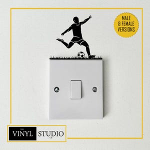 Op de afbeelding: Een witte lichtschakelaar met een zwarte vinylsticker van een voetballer die een bal trapt boven een strook gras. Een gele cirkelvormige badge rechtsboven bevat de tekst MALE & FEMALE VERSIONS in zwarte hoofdletters.