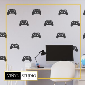 Könnte beinhalten: Eine weiße Wand, die mit mehreren dunkelgrauen Aufklebern von Videospiel-Controllern verziert ist. Im Vordergrund befindet sich ein Schreibtisch mit Computer, Lampe und Stuhl. Das Logo Vinyl Studio GB befindet sich in der unteren linken Ecke.