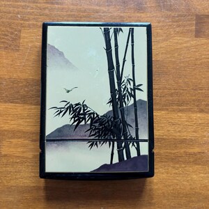 May include: A black rectangular box with a hinged lid. The lid features a serene landscape painting with bamboo stalks, mountains, and a flying bird. The artwork is in shades of black, white, and purple.