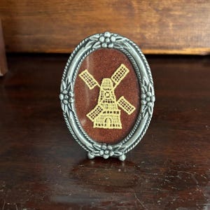 May include: An oval-shaped silver-toned metal frame featuring a floral relief pattern along the border. Inside the frame, a dark brown fabric backing displays a light yellow embroidered windmill design with four blades and a central structure.