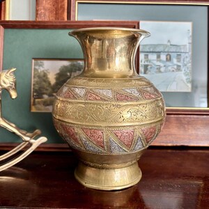 May include: A vintage brass vase with detailed etched patterns and a flared rim. The vase features bands of detailed designs in gold, copper, and silver tones. The base is a pedestal shape, and the overall aesthetic is ornate and decorative.