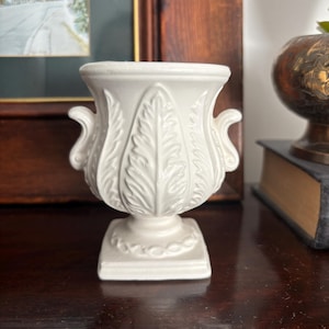 May include: A white ceramic urn-style vase featuring a prominent embossed leaf pattern on the exterior, two curved side handles, and a square base. The vessel has a wide, flared rim and a smooth, matte finish, showcasing a classic, symmetrical design.