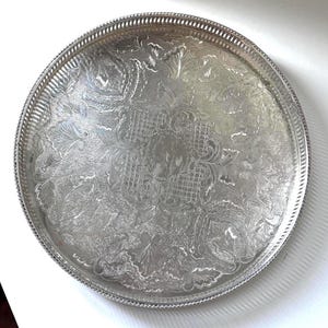 May include: A round silver-coloured metal serving tray featuring an intricate embossed floral and lattice pattern across the surface. The tray has a raised, decorative pierced gallery rim with a beaded edge, providing a textured metallic finish.