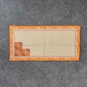 May include: Beige rectangular table runner with an orange embroidered border and corner designs. The border has a repeating geometric pattern. The corner designs are orange and brown. The runner is made of woven fabric.