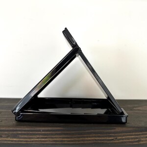 May include: A black, open, rectangular compact case with a hinged lid. The case is positioned at an angle, revealing the interior. The surface is glossy, reflecting light. The background is a white wall and a dark wooden surface.