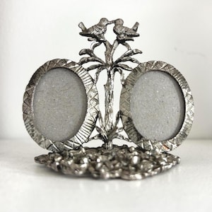 May include: A silver-toned metal double photo frame featuring two oval-shaped openings with textured, cross-hatched borders. The frame is supported by a central tree-like structure topped with two birds facing each other, resting on a circular base.