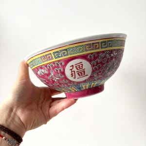 May include: A decorative ceramic bowl with a vibrant pink background and intricate floral patterns. The bowl features a white circle with Chinese characters. The rim has a yellow and blue geometric design.