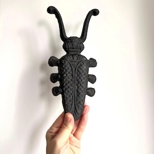 May include: A black cast iron decorative bug-shaped object. The object has a detailed design with a textured body, six legs, and two curved antennae. The bug is held in a hand against a white background.