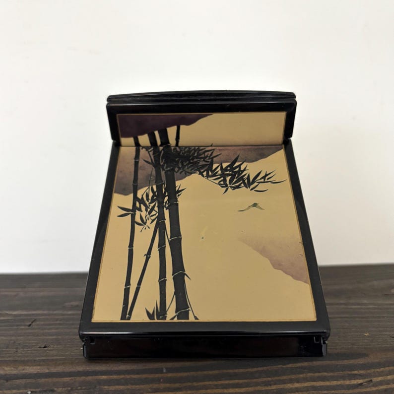 May include: Black rectangular box with a hinged lid. The interior features a bamboo forest scene in black and tan tones. The box is set on a dark wooden surface.