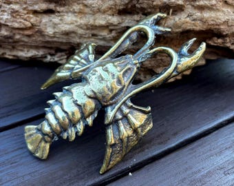 May include: A metallic brass-toned lobster figurine featuring a segmented tail, detailed claws, and curved antennae. The surface of the crustacean sculpture displays a weathered, antique finish with visible texture and highlights across its body.
