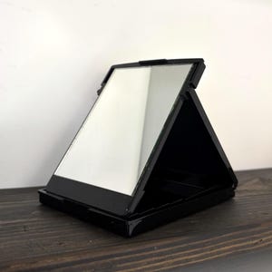 May include: A black, rectangular, folding compact mirror. The mirror is open, revealing a reflective surface. The compact is positioned on a dark wooden surface against a white background. The mirror is angled, showing its reflective surface.