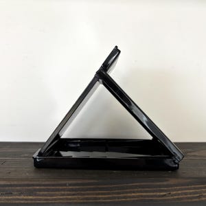 May include: A black plastic case, open to form a triangular shape. The case is on a dark wooden surface, with a white wall in the background. The case appears to be empty.