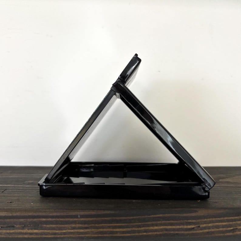 May include: A black plastic case, open to form a triangular shape. The case is on a dark wooden surface, with a white wall in the background. The case appears to be empty.