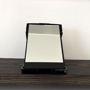 May include: A black rectangular compact mirror with a silver reflective surface. The mirror is open, revealing the reflective surface. The compact is sitting on a dark brown wooden surface.