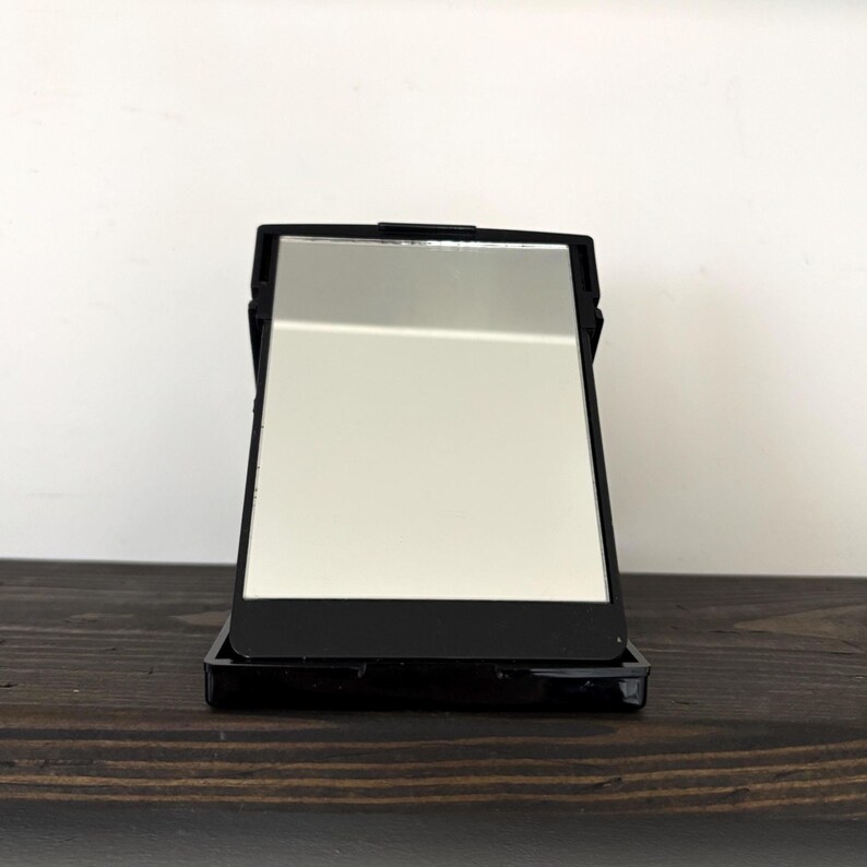 May include: A black rectangular compact mirror with a silver reflective surface. The mirror is open, revealing the reflective surface. The compact is sitting on a dark brown wooden surface.