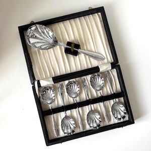 May include: A set of silver-colored serving spoons and utensils in a black box with a cream-colored interior. The spoons have a shell-like design on the bowl. The box is open, revealing the spoons and utensils arranged neatly inside.