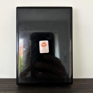 May include: A rectangular, black lacquered box with a glossy finish. A small, rectangular sticker with red and white text and a logo is affixed to the front. The sticker reads "TAKE JAPAN ART COLLECTION".