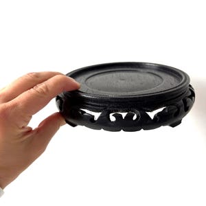 May include: A round, black wooden display stand with a carved, openwork design around the base. The stand has a flat top surface and is designed to hold a decorative object. The stand is held by a hand against a white background.