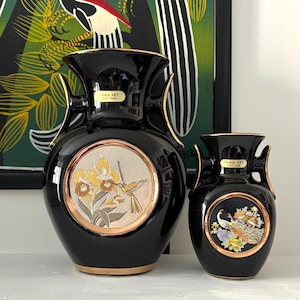 May include: Two black ceramic vases with gold trim and Chokin ART 24KT Gold labels. The larger vase features a circular medallion with a hummingbird and yellow flowers. The smaller vase has a circular medallion depicting a peacock and flowers.