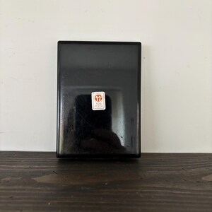May include: A rectangular, black box with a glossy finish. A small, white and red sticker is affixed to the front. The box is set against a white wall and a dark wooden surface.