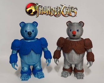 Puede incluir: Dos figuras de ThunderCats impresas en 3D, una azul y otra plateada, ambas con un diseño de oso y narices rojas. Las figuras están de pie sobre una superficie blanca con el logotipo de ThunderCats en el fondo.