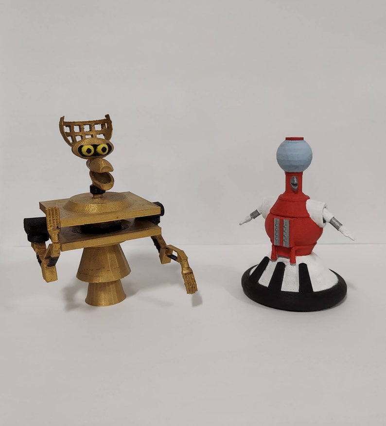 May include: Two robot figurines, one gold with a round base and a square platform, the other red with a white base and a round top.