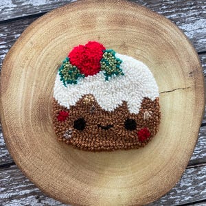 May include: A brown and white Christmas pudding with a red and green holly sprig on top. The pudding has a cute cartoon face with black eyes and a smile.