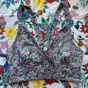 Puede incluir: Un top corto con estampado floral y diseño de tirantes cruzados. El top presenta una mezcla de flores moradas, verdes y blancas sobre un fondo oscuro. Las correas se cruzan en la espalda y la tela tiene una apariencia texturizada. El top es una prenda elegante.