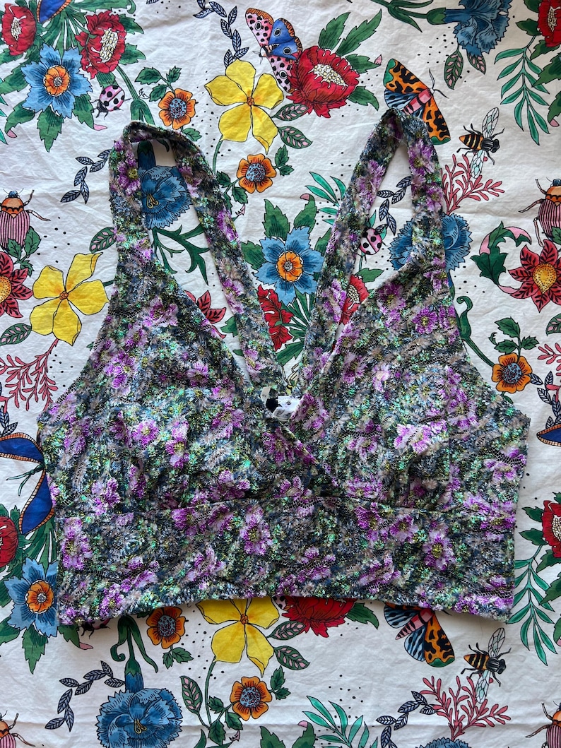 May include: A floral print crop top with a cross-strap design. The top features a mix of purple, green, and white flowers on a dark background. The straps cross in the back, and the fabric has a textured appearance. The top is a stylish piece of apparel.