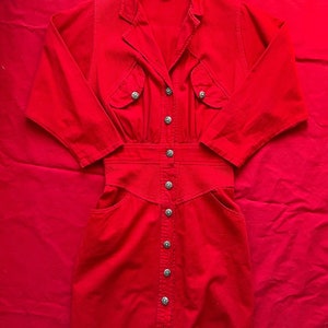 May include: A red corduroy dress with a button-up front and long sleeves. The dress has a fitted waist and a flared skirt. The dress has a V-neckline and a collar. The dress has pockets on the front and a label that says "Versace".