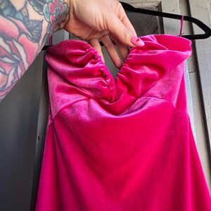 Puede incluir: Un vibrante vestido fucsia sin tirantes hecho de suave tejido de terciopelo. El corpi&ntilde;o presenta un escote coraz&oacute;n fruncido. El vestido est&aacute; colgado de una percha negra.