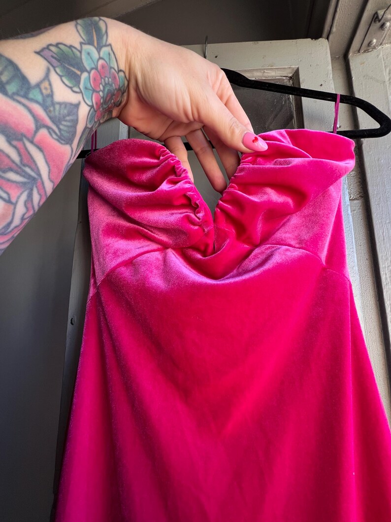 Puede incluir: Un vibrante vestido fucsia sin tirantes hecho de suave tejido de terciopelo. El corpi&ntilde;o presenta un escote coraz&oacute;n fruncido. El vestido est&aacute; colgado de una percha negra.