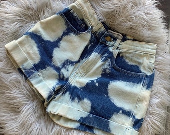 May include: Blue denim shorts with a white bleach tie-dye pattern. The shorts have a cuffed hem and a label that reads "American Apparel".