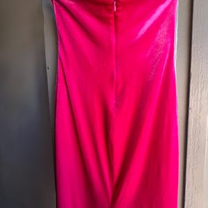 Puede incluir: Un vibrante vestido palabra de honor fucsia, confeccionado con un material suave y aterciopelado. El vestido est&aacute; colgado de una percha negra, mostrando su dise&ntilde;o elegante y ajustado. Una cremallera recorre la espalda, a&ntilde;adiendo un toque de detalle a la elegante prenda.