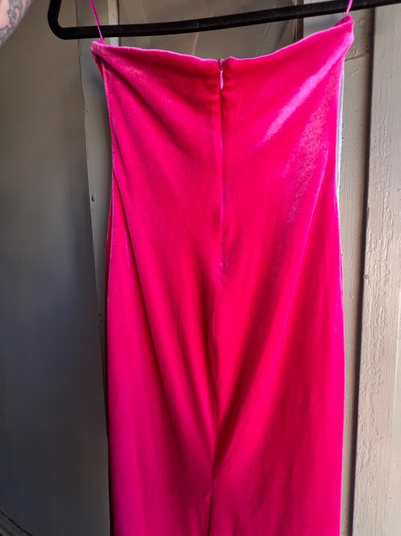 Puede incluir: Un vibrante vestido palabra de honor fucsia, confeccionado con un material suave y aterciopelado. El vestido est&aacute; colgado de una percha negra, mostrando su dise&ntilde;o elegante y ajustado. Una cremallera recorre la espalda, a&ntilde;adiendo un toque de detalle a la elegante prenda.