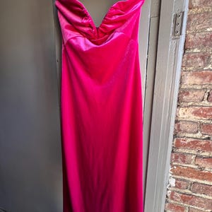 Puede incluir: Un vibrante vestido fucsia sin tirantes cuelga de una percha negra. El vestido presenta un escote coraz&oacute;n con tela fruncida y un dise&ntilde;o elegante hasta el suelo. El vestido est&aacute; hecho de un material brillante y sedoso.