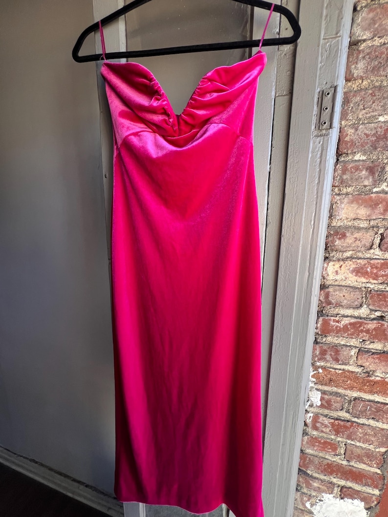 Puede incluir: Un vibrante vestido fucsia sin tirantes cuelga de una percha negra. El vestido presenta un escote coraz&oacute;n con tela fruncida y un dise&ntilde;o elegante hasta el suelo. El vestido est&aacute; hecho de un material brillante y sedoso.