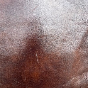 May include: Close-up of a brown leather surface with a smooth, shiny finish. The leather has a subtle texture and a few light scratches.