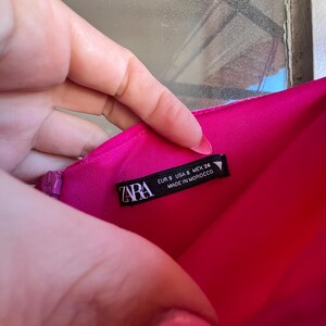 Puede incluir: Una prenda rosa brillante con una etiqueta que dice "ZARA" e incluye informaci&oacute;n de tallas: EUR S, USA S, MEX 26. La etiqueta tambi&eacute;n indica "MADE IN MOROCCO". La prenda es sostenida por una mano.