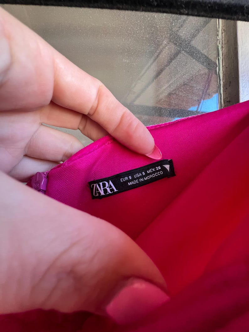 Puede incluir: Una prenda rosa brillante con una etiqueta que dice "ZARA" e incluye informaci&oacute;n de tallas: EUR S, USA S, MEX 26. La etiqueta tambi&eacute;n indica "MADE IN MOROCCO". La prenda es sostenida por una mano.