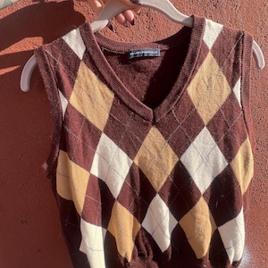 Puede incluir: Un chaleco de punto marrón con diseño argyle y cuello en V. El chaleco presenta un patrón de rombos en colores marrón, beige y crema. El chaleco se muestra en una percha.