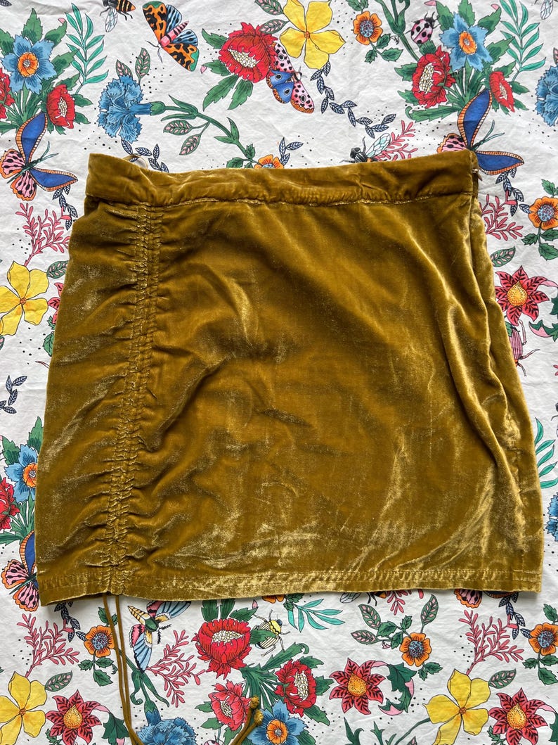 Op de afbeelding: Een mosterdgele mini-rok van fluweel met een trekkoord in de taille en een geplooide zijkant.