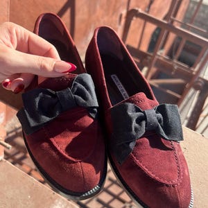 Puede incluir: Un par de mocasines de ante burdeos con lazos negros. Los zapatos tienen una puntera redondeada y una suela negra. El nombre de la marca "MANOLO BLAHNIK" es visible en la plantilla. Los mocasines se exhiben sobre una superficie de madera.