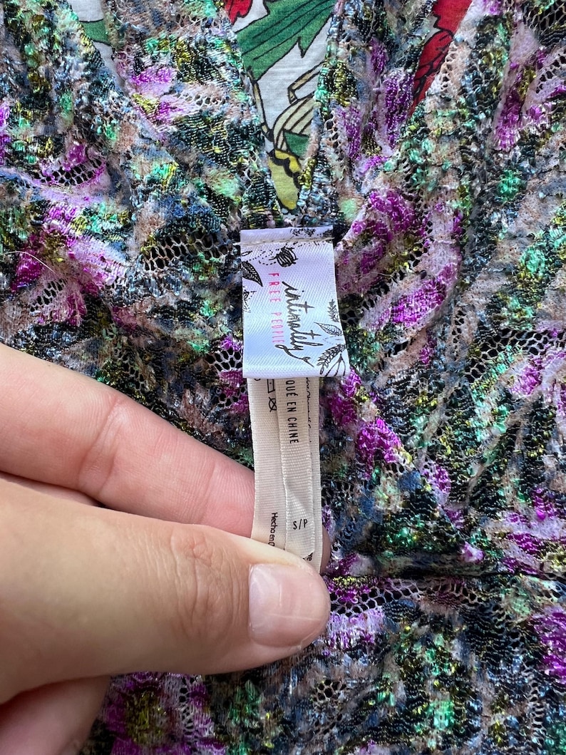 May include: Close-up of a floral patterned garment with a V-neck. The fabric is a mix of purple, green, and gold. A tag reads "intimately FREE PEOPLE" and "QUE EN CHINE". The garment is size S/P.